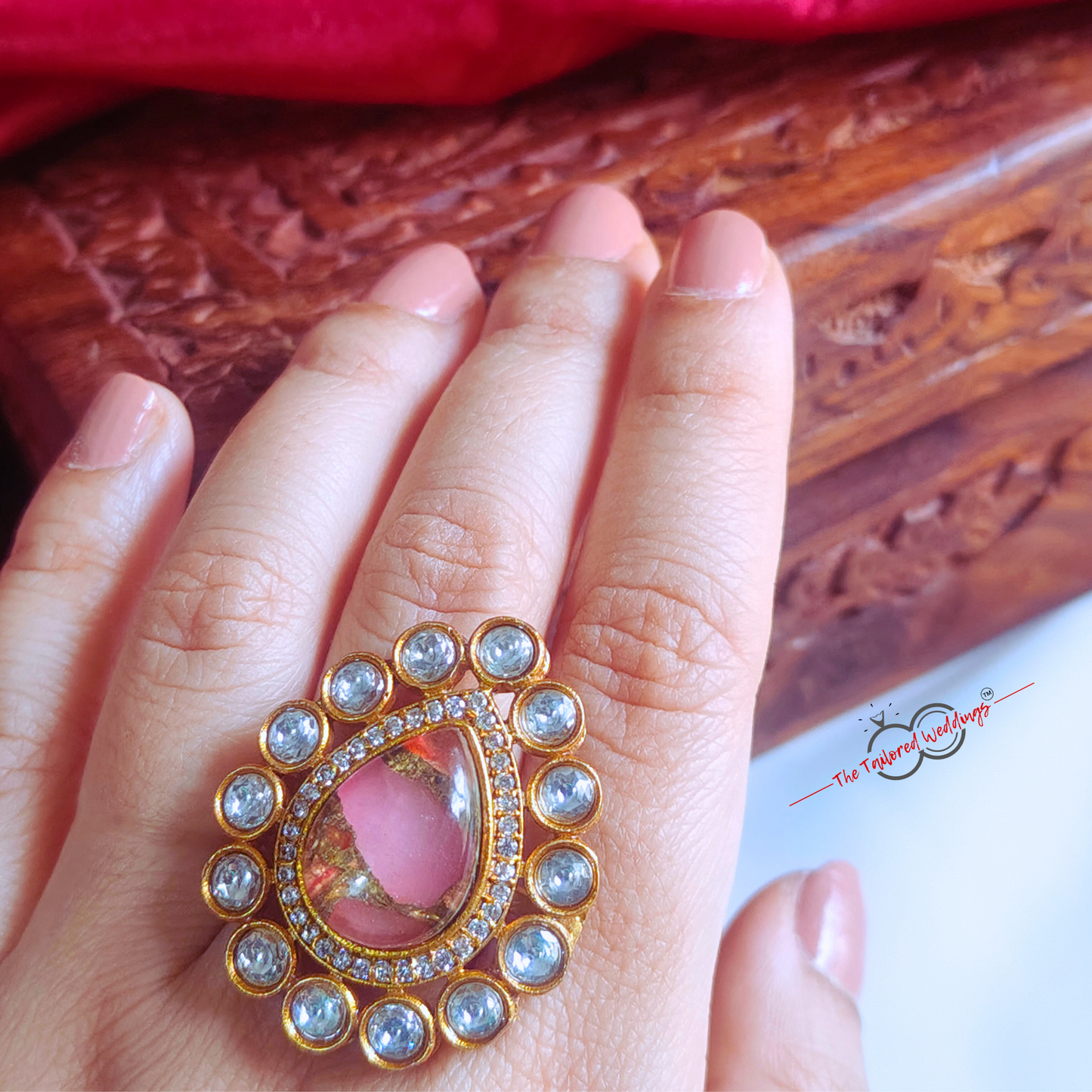 Decorative ring with pink gem and gold accents on a white surface, with a wooden box in the background.