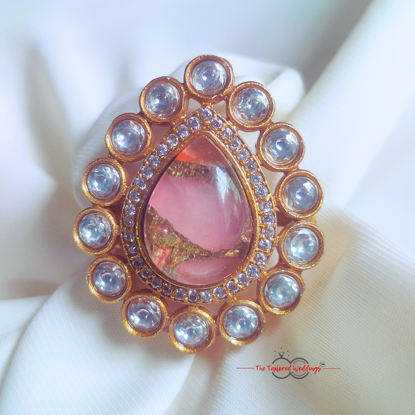 Decorative ring with pink gem and gold accents on a white surface, with a wooden box in the background.