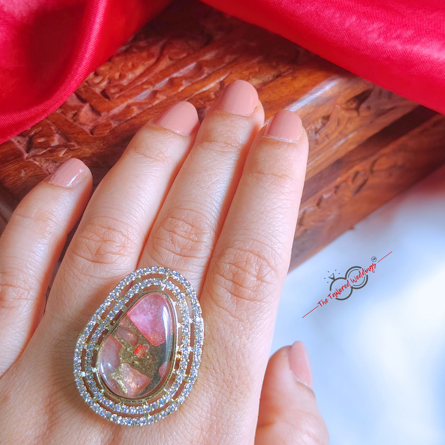 Decorative ring with pink gem and gold accents on a white surface, with a wooden box in the background.
