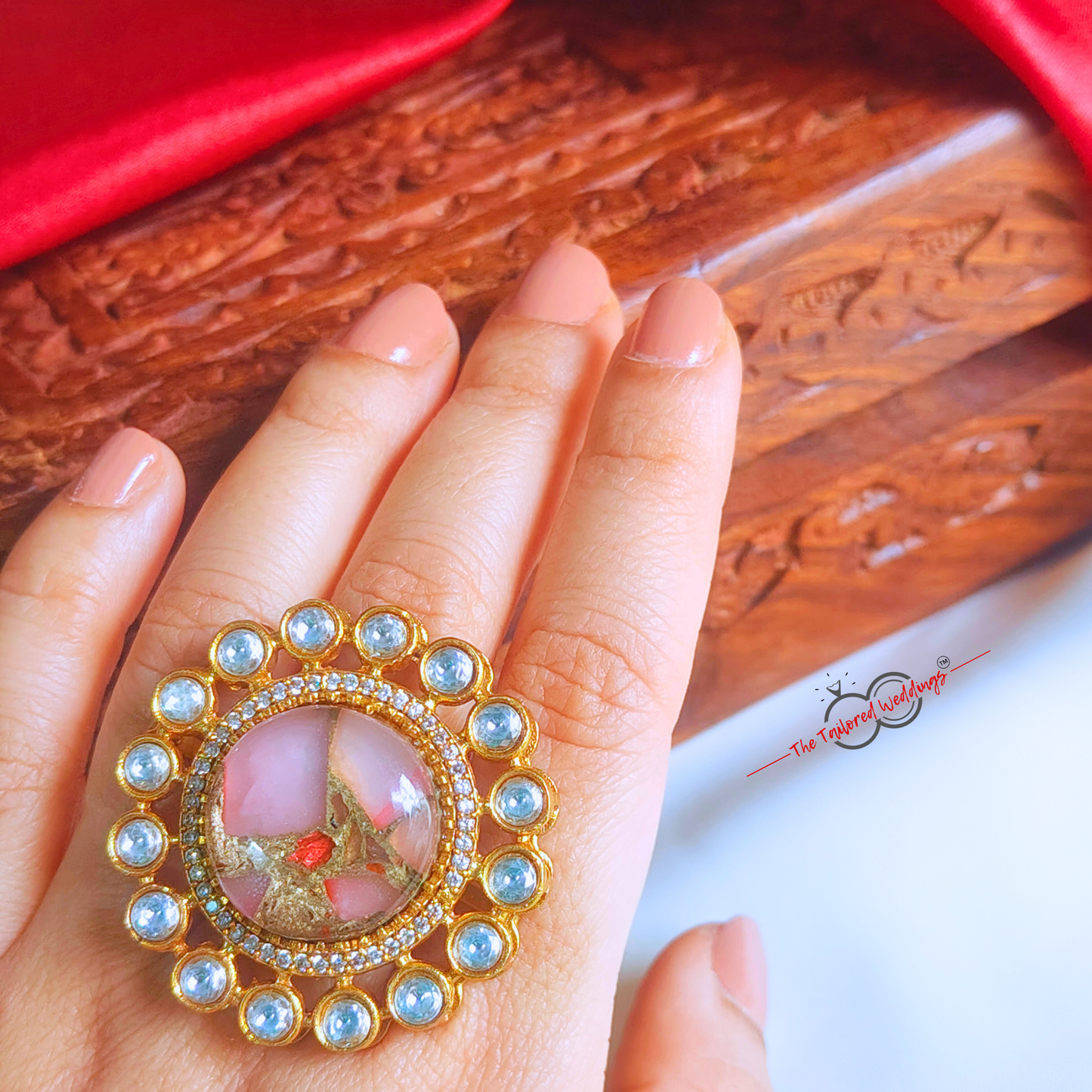 Decorative ring with rose pink gemstone in the centre with small circle encircles the centre piece, placed on a white surface next to a wooden box