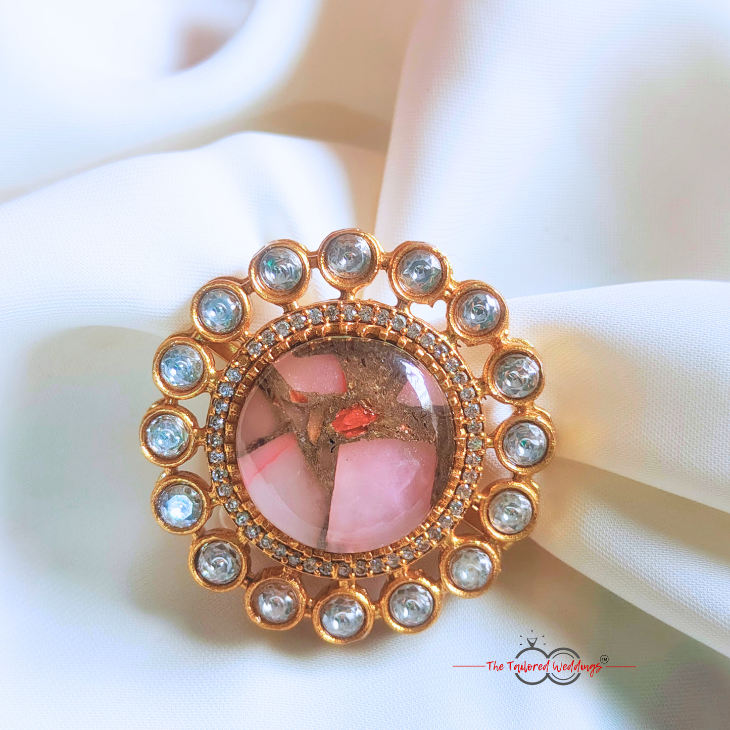 Decorative ring with rose pink gemstone in the centre with small circle encircles the centre piece, placed on a white surface next to a wooden box