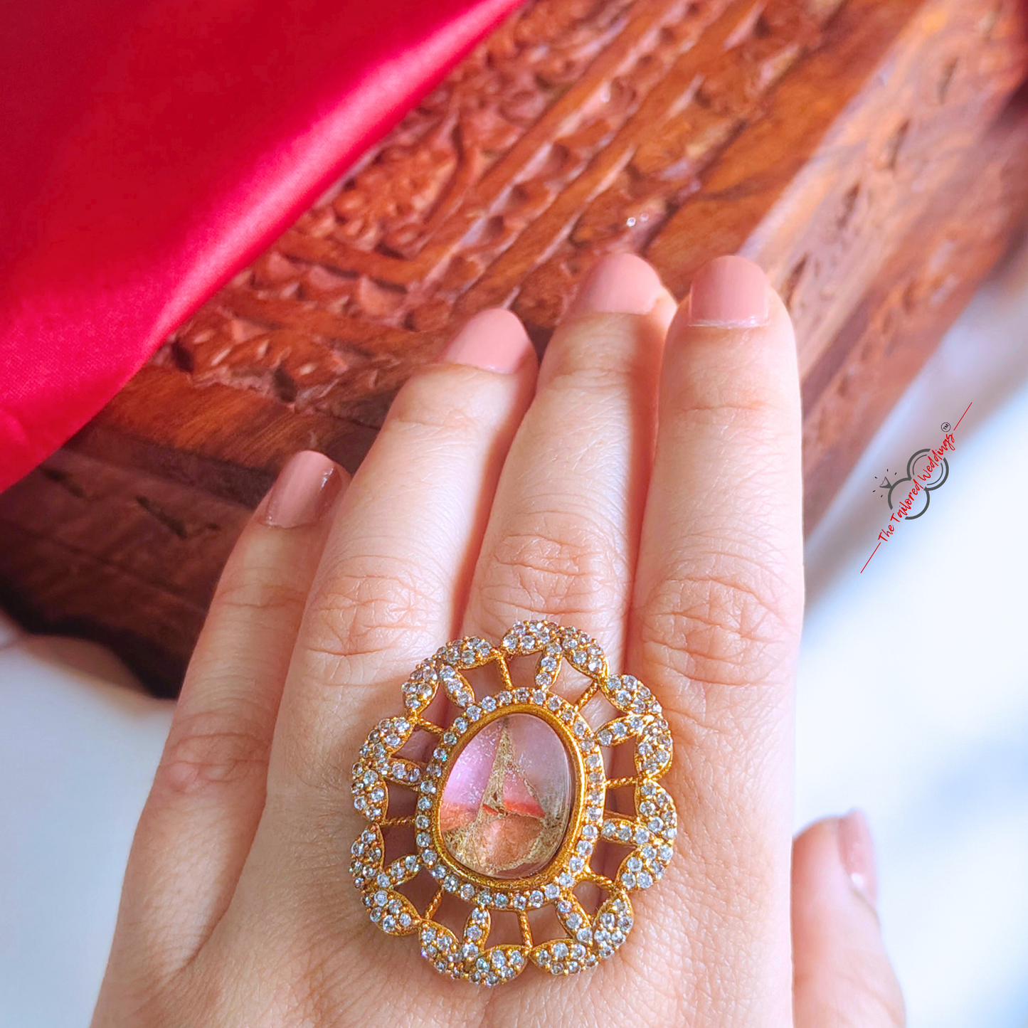 Gold toned ring with a pink stone on a white surface with a wooden box in the background