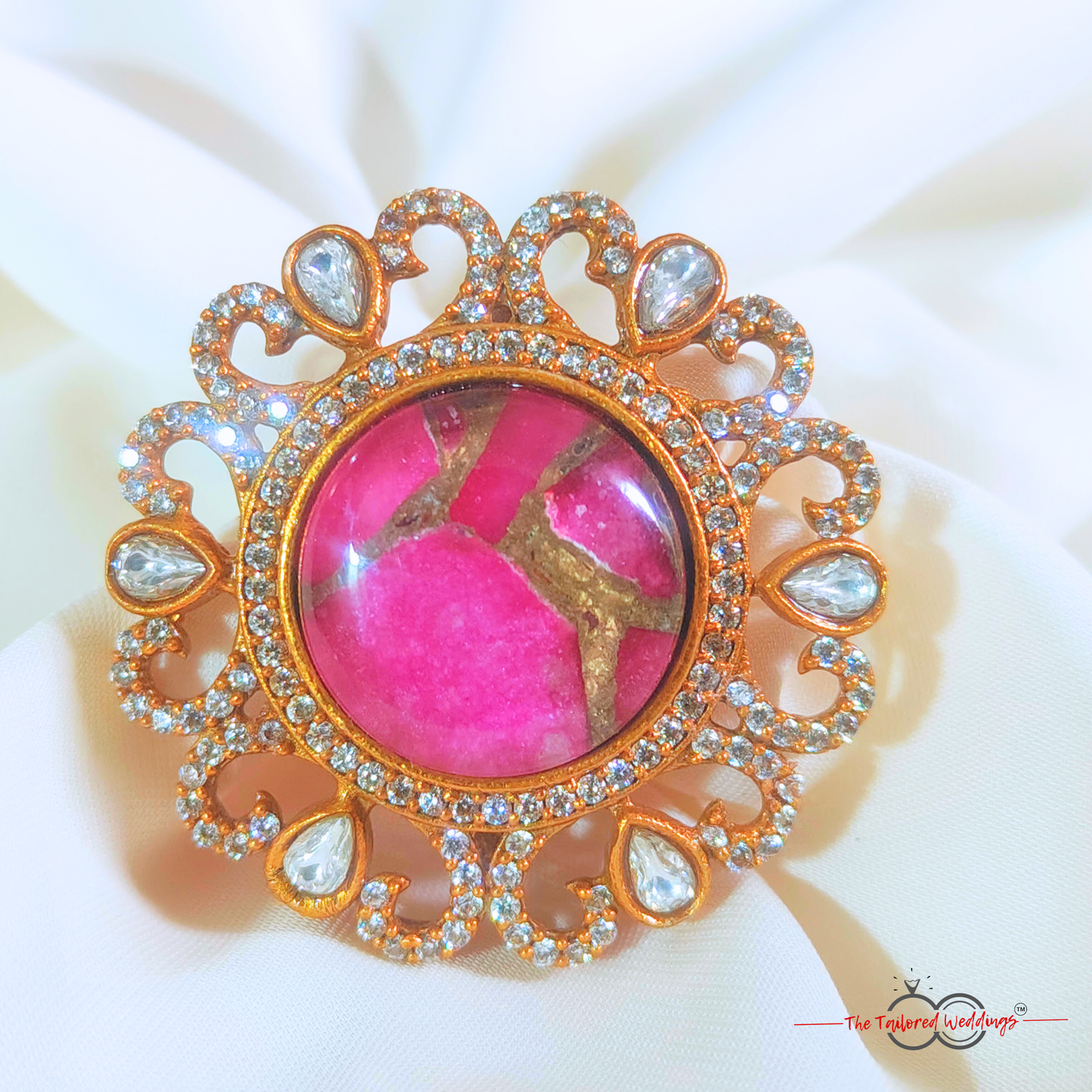 Decorative ring with magenta pink gem stone at the centre and gold accents around it, placed on a white surface next to a wooden box