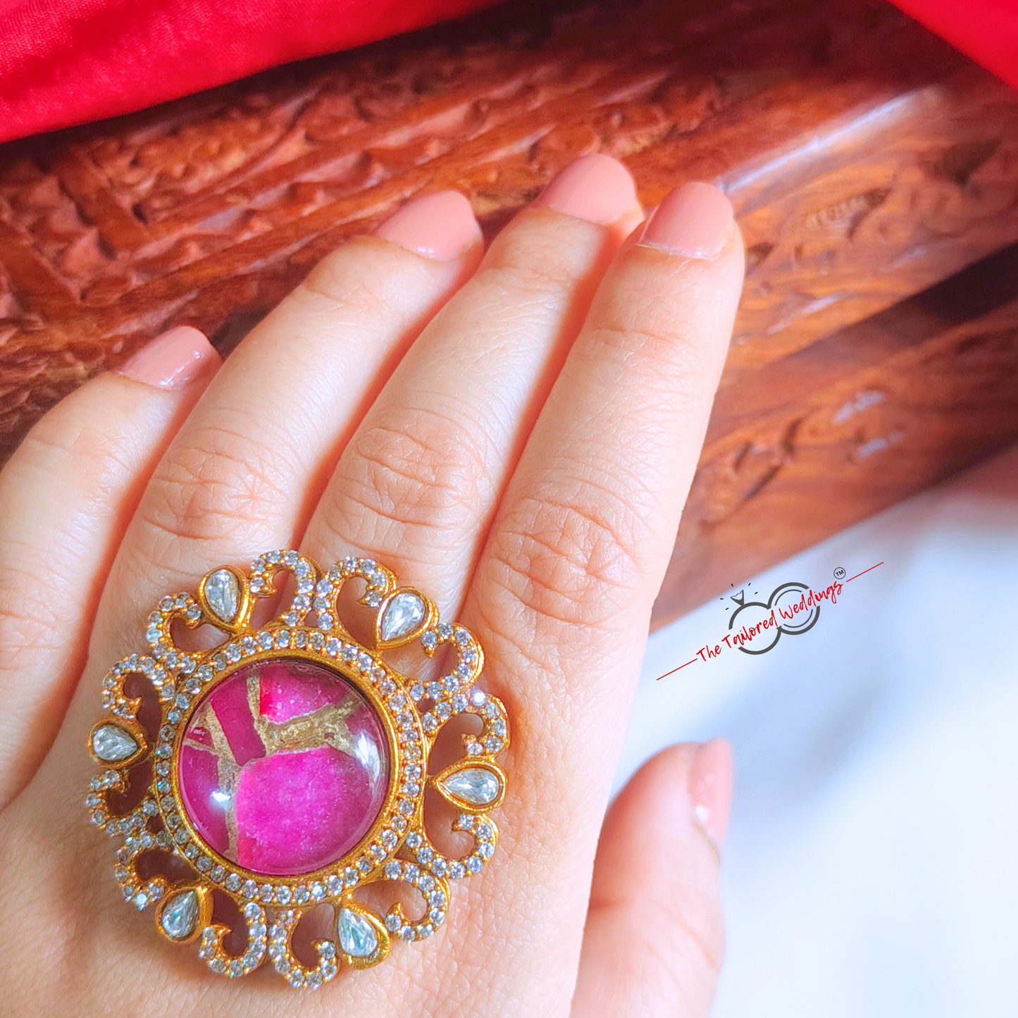 Decorative ring with magenta pink gem stone at the centre and gold accents around it, placed on a white surface next to a wooden box