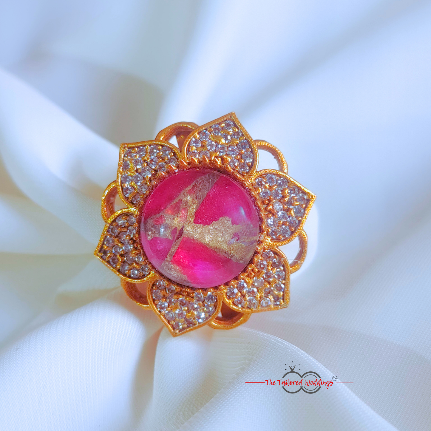 Decorative ring with pink gem and gold accents on a white surface, with a wooden box in the background.