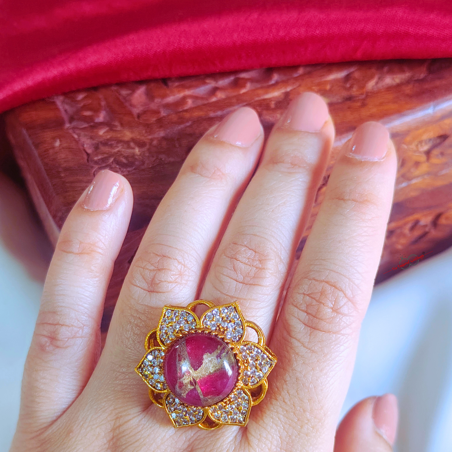 Decorative ring with pink gem and gold accents on a white surface, with a wooden box in the background.