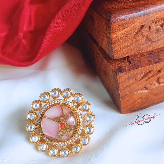 Decorative ring with rose pink gemstone in the centre with small circle encircles the centre piece, placed on a white surface next to a wooden box