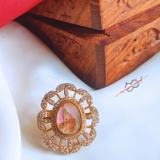 Gold toned ring with a pink stone on a white surface with a wooden box in the background