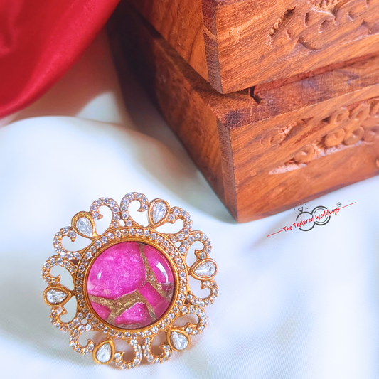 Decorative ring with magenta pink gem stone at the centre and gold accents around it, placed on a white surface next to a wooden box