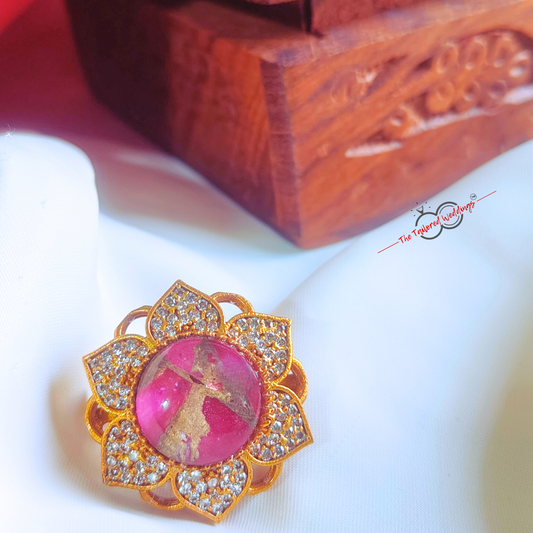 Decorative ring with pink gem and gold accents on a white surface, with a wooden box in the background.